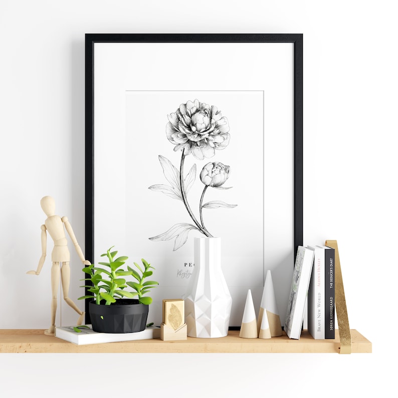 Op de afbeelding: Een zwart ingelijst afdruk van een zwart-witte tekening van een pioenroos, een witte geometrische vaas, een kleine plant in een pot, een houten mannequin en een stapel boeken op een houten plank.