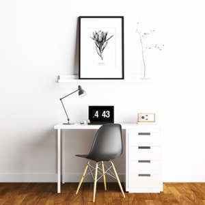 May include: A white desk with a black lamp, a laptop, a clock, and a white drawer unit. A black framed print of a black and white drawing of a flower hangs above the desk. A small branch with red berries is in a vase on the shelf above the desk.