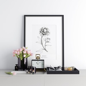 Op de afbeelding: Een zwart ingelijste print van een zwart-witte tekening van een pioenroos met de tekst "PEONY Paeonia Lactiflora" onder de afbeelding. De print staat op een witte plank met een zwarte dienblad, een klok, kaarsen, een vaas met roze bloemen en een boek.