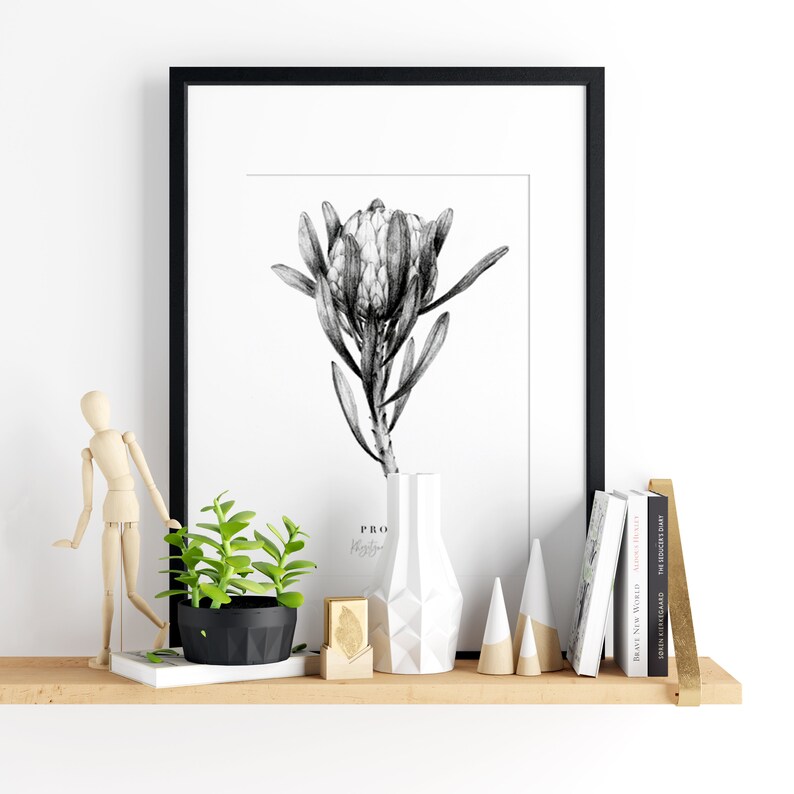 May include: A black and white framed print of a protea flower, a wooden shelf with a wooden mannequin, a potted succulent, a gold leaf, a white geometric vase, and a stack of books with gold accents.