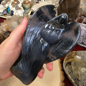 Puede incluir: Una figura de ángel gris oscuro tallada en piedra. El ángel tiene las alas extendidas y una expresión serena. La escultura se sostiene en una mano, mostrando la artesanía detallada y la superficie lisa y pulida. La figura es un objeto decorativo.
