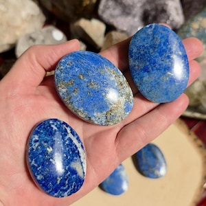 May include: Several oval-shaped lapis lazuli stones in varying shades of blue, flecked with gold and white. The stones are smooth and polished, showcasing natural patterns. The stones are held in a hand.