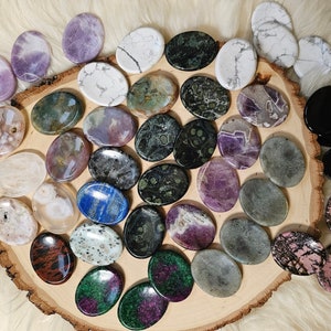 May include: A collection of polished oval-shaped stones in various colors and patterns, including purple, green, black, white, and brown. The stones are arranged on a wooden slice.