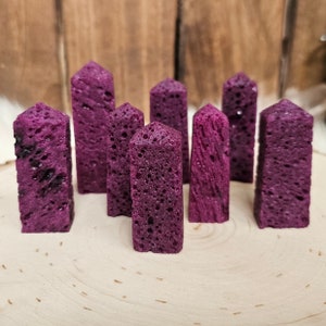 May include: A collection of eight upright, rectangular, purple-colored stone obelisks with textured surfaces. The obelisks are arranged on a light-colored wooden surface, with a blurred wooden background.