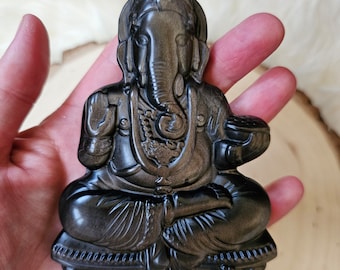 Op de afbeelding: Een zwart en gouden Ganesha-beeld, een hindoeïstische godheid, zittend in een meditatieve houding met een slurf en grote oren. Het beeld is gemaakt van gepolijst obsidiaan en heeft een glanzend, reflecterend oppervlak.