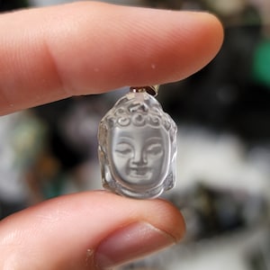 Puede incluir: Un colgante de cuarzo transparente tallado con un rostro de Buda detallado. El colgante es de color gris claro y tiene un acabado liso y pulido.