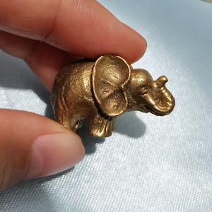 Puede incluir: Una pequeña figurita de elefante de color dorado. El elefante está de pie sobre sus cuatro patas y tiene la trompa levantada. La figurita está hecha de metal y tiene un diseño detallado.