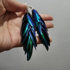 Puede incluir: Un par de pendientes largos e iridiscentes hechos con alas de escarabajo. Las alas están superpuestas y tienen una combinación de colores azul, verde y negro.