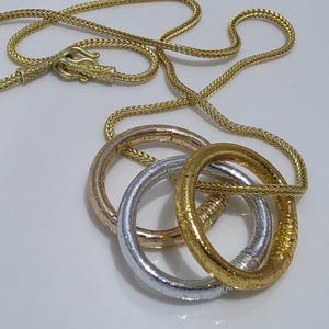 Op de afbeelding: Een goudkleurige tarweketting met drie in elkaar grijpende ronde hangers. De hangers hebben een getextureerde, metallic afwerking in de tinten roségoud, zilver en geelgoud. De ketting is voorzien van een veilige, versierde haaksluiting.