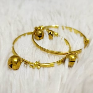 Peut inclure: Deux bracelets en or avec de petites cloches attachées. Les bracelets sont sur une surface blanche et duveteuse.