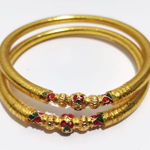 Puede incluir: Dos brazaletes dorados con superficies texturizadas. Cada brazalete presenta una sección decorativa con detalles rojos y verdes. Los brazaletes están apilados, creando un efecto en capas, sobre un fondo blanco.