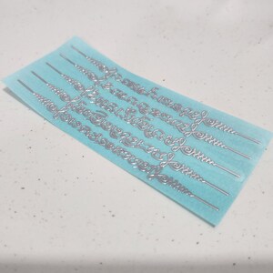 May include: A sheet of silver-colored decorative stickers on a light blue background. The stickers are in the shape of a repeating pattern of swirls.