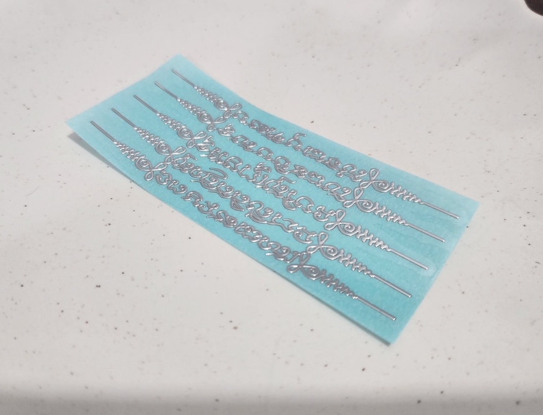 May include: A sheet of silver-colored decorative stickers on a light blue background. The stickers are in the shape of a repeating pattern of swirls.