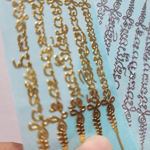 May include: Five gold and silver decorative stickers with intricate designs and lettering on a light blue background.