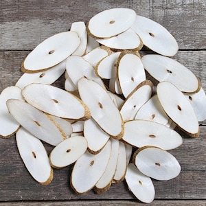 Könnte beinhalten: Ein Stapel weißer ovaler Holzscheiben mit einem Loch in der Mitte. Die Holzscheiben sind in einem zufälligen Muster angeordnet und haben eine natürliche Holzmaserung.