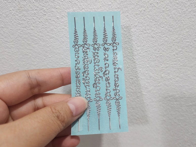 May include: Four silver temporary tattoos on a light blue background. The tattoos feature intricate designs and text in the Thai language.