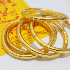 May include: A set of four gold-colored bangles with intricate designs. The bangles are stacked on top of each other and are resting on a yellow surface with Thai text.