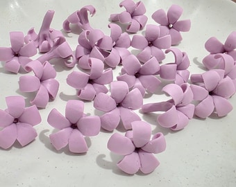 May include: A collection of light purple, handmade flower embellishments. The flowers have five petals and are arranged on a white surface. Some flowers have stems, and all have a soft, rounded appearance.