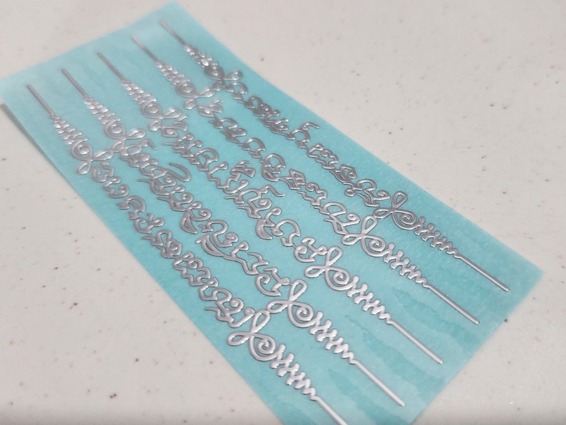 May include: A sheet of silver-colored decorative stickers with intricate designs. The stickers are on a light blue background.