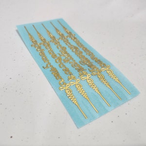 May include: A sheet of gold-colored stickers with intricate designs. The stickers are on a light blue background and are arranged in rows. Each sticker is a long, thin strip with a repeating pattern of swirls and lines.
