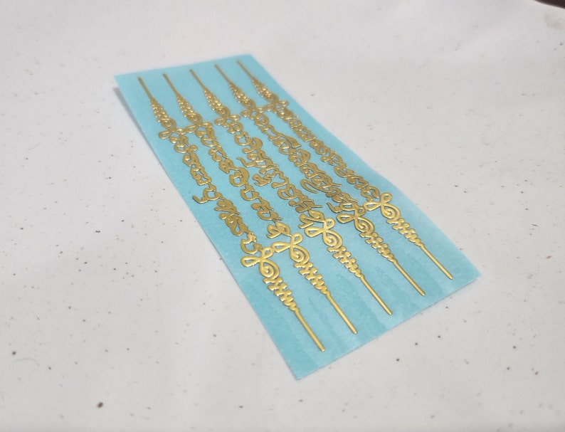 May include: A sheet of gold-colored stickers with intricate designs. The stickers are on a light blue background and are arranged in rows. Each sticker is a long, thin strip with a repeating pattern of swirls and lines.