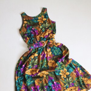 May include: A sleeveless maxi dress with a floral print in shades of yellow, green, purple, and brown. The dress has a fitted bodice and a flowing skirt. It features a gold belt with a buckle.