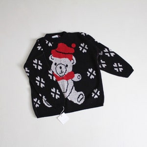 May include: Black knitted sweater with a white and gray bear wearing a red Santa hat and a red scarf. The sweater has a snowflake pattern.