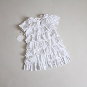 May include: White ruffled dress with short sleeves. The dress has multiple layers of ruffles and a simple neckline.