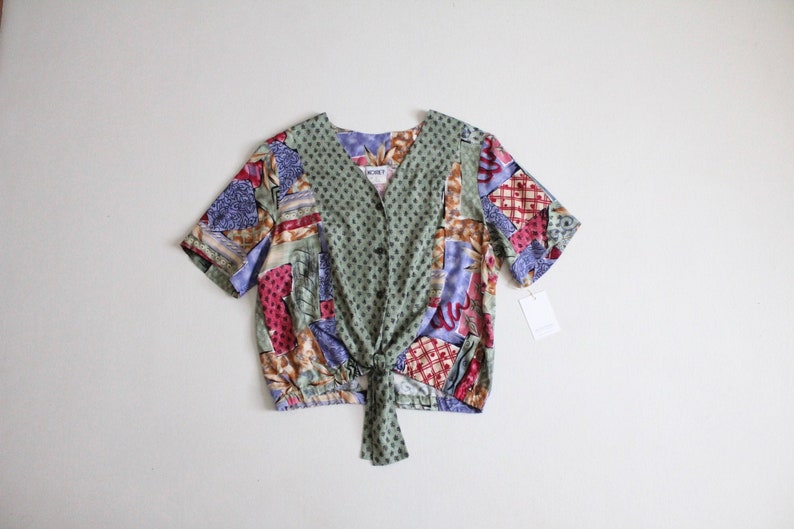May include: A green, blue, red, and brown patterned short-sleeved blouse with a tie front and button closure. The blouse has a v-neck and is made of a silky fabric.