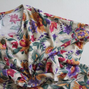May include: A cream-colored button-up shirt with a floral print featuring shades of red, purple, yellow, and green. The shirt has a V-neck and short sleeves. The label reads "Van Otis".