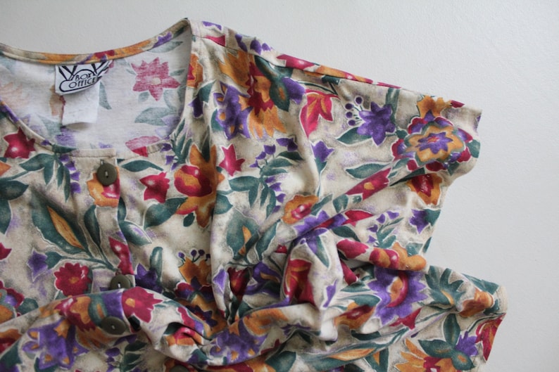 May include: A cream-colored button-up shirt with a floral print featuring shades of red, purple, yellow, and green. The shirt has a V-neck and short sleeves. The label reads "Van Otis".