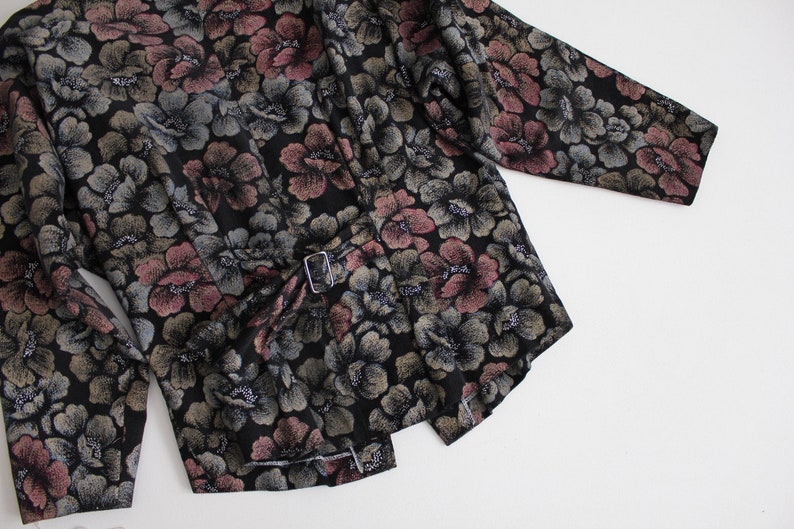 May include: A black long-sleeved jacket with a floral print in shades of pink, green, and silver. The jacket has a belt with a silver buckle.