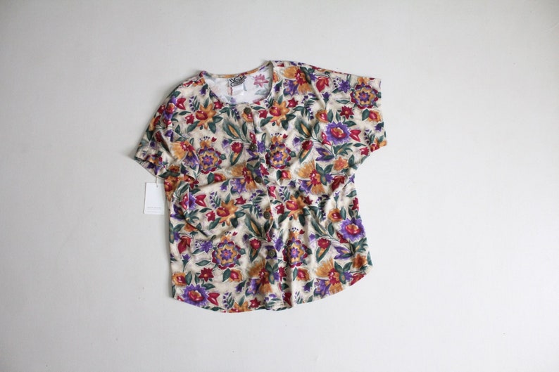 May include: A short-sleeved, cream-colored button-up shirt with a floral print featuring red, yellow, purple, and green flowers.