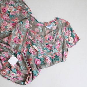 Op de afbeelding: Een vintage bloemenjurk in roze, groene en witte tinten op een grijze achtergrond. De jurk heeft een V-hals, korte mouwen en een zwierige rok. Er is een blanco label aan bevestigd.