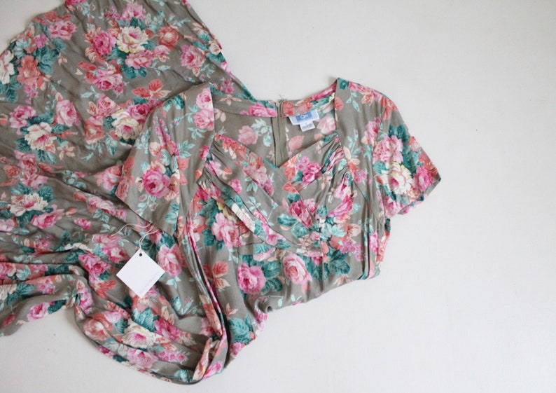 Op de afbeelding: Een vintage bloemenjurk in roze, groene en witte tinten op een grijze achtergrond. De jurk heeft een V-hals, korte mouwen en een zwierige rok. Er is een blanco label aan bevestigd.