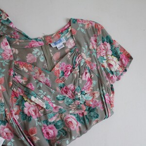 Op de afbeelding: Een vintage jurk met een bloemenpatroon van roze rozen en groene bladeren op een grijze achtergrond. De jurk heeft een V-hals, korte mouwen en een ritssluiting. Er is een label aan de jurk bevestigd.