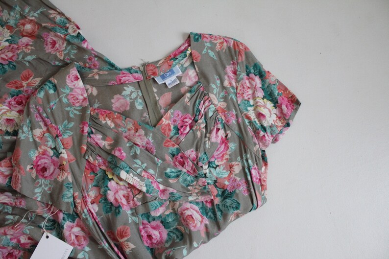 Op de afbeelding: Een vintage jurk met een bloemenpatroon van roze rozen en groene bladeren op een grijze achtergrond. De jurk heeft een V-hals, korte mouwen en een ritssluiting. Er is een label aan de jurk bevestigd.