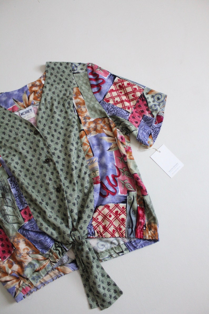May include: A green, purple, red, and brown patterned shirt with a tie front and short sleeves. The shirt has a button closure and the label "KOret" is visible.
