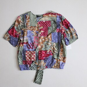 May include: A short-sleeved, tie-front blouse with a patchwork design featuring various shades of green, blue, red, and brown. The fabric has a floral and geometric pattern.