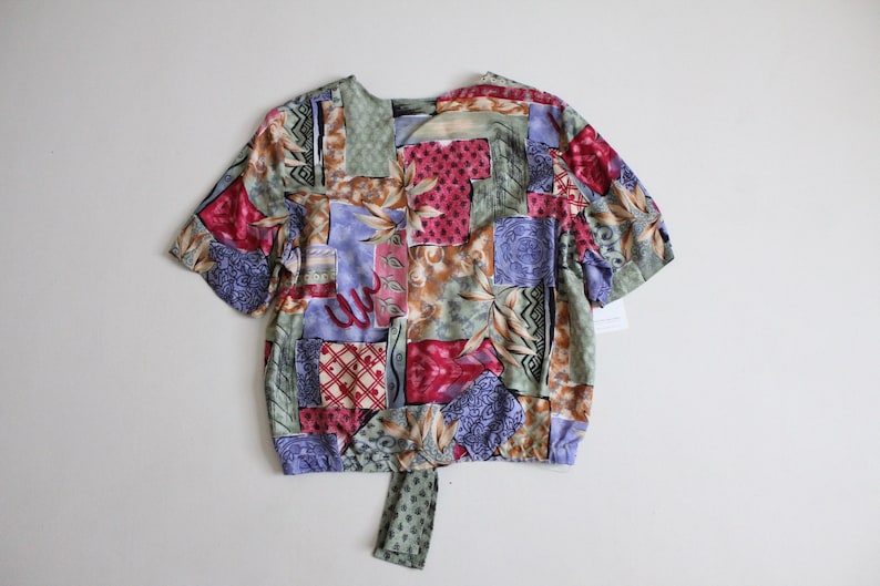 May include: A short-sleeved, tie-front blouse with a patchwork design featuring various shades of green, blue, red, and brown. The fabric has a floral and geometric pattern.