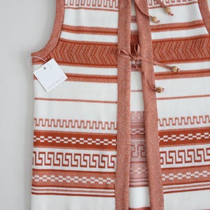 Op de afbeelding: Een wit en oranje gestreepte gebreide vest met een strik sluiting en houten kralen. De vest heeft een geometrisch patroon en het label staat "KNIT PRETTY".
