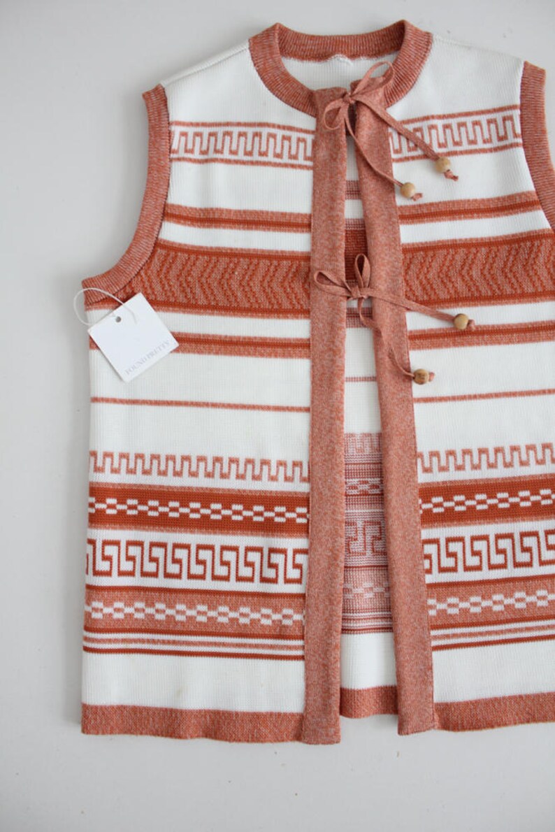 Op de afbeelding: Een wit en oranje gestreepte gebreide vest met een strik sluiting en houten kralen. De vest heeft een geometrisch patroon en het label staat "KNIT PRETTY".
