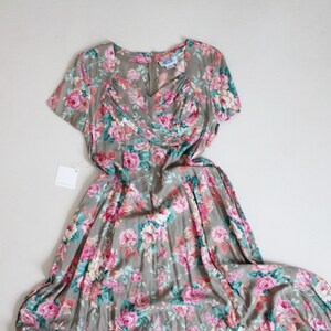 Op de afbeelding: Vintage jurk met bloemenpatroon van roze en rode rozen op een grijze achtergrond. De jurk heeft korte mouwen, een V-hals en een getailleerd lijfje. Er is een label bevestigd. Klassieke stijl.