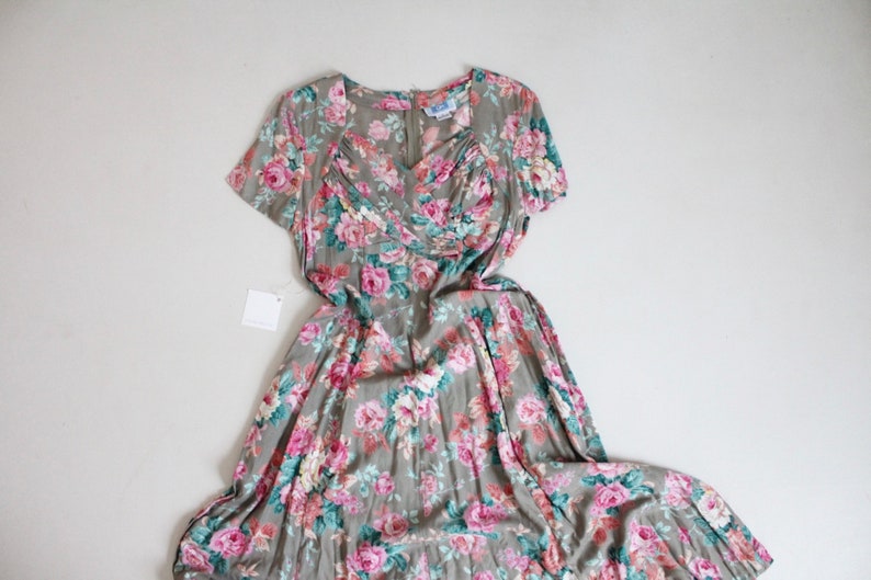 Op de afbeelding: Vintage jurk met bloemenpatroon van roze en rode rozen op een grijze achtergrond. De jurk heeft korte mouwen, een V-hals en een getailleerd lijfje. Er is een label bevestigd. Klassieke stijl.