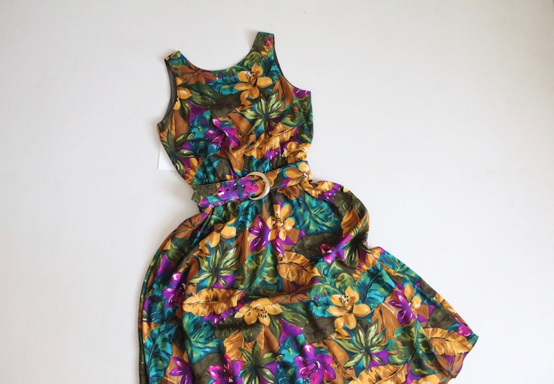 May include: A sleeveless maxi dress with a floral print in shades of yellow, green, purple, and brown. The dress has a fitted bodice and a flowing skirt. It features a gold belt with a buckle.