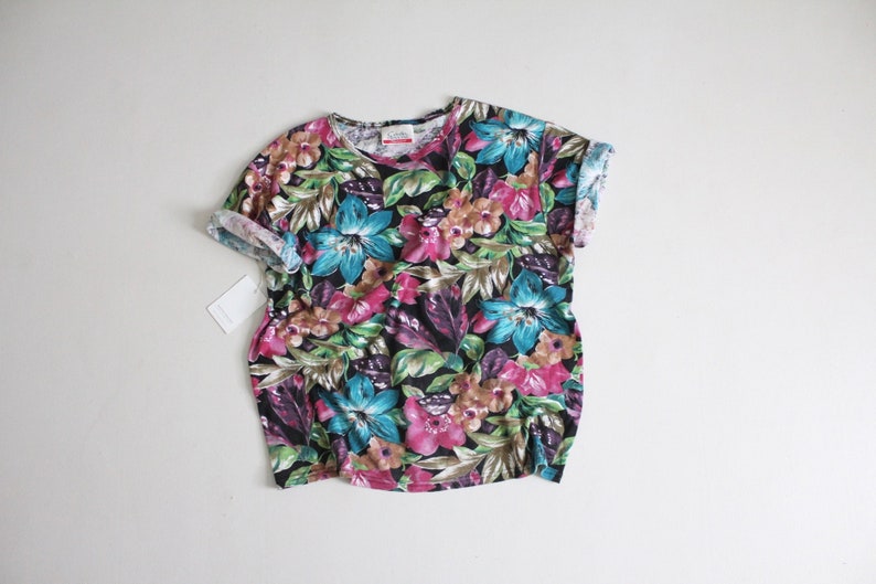 May include: A short-sleeved t-shirt with a tropical floral print in shades of pink, blue, green, and brown. The shirt has a crew neck and a relaxed fit.
