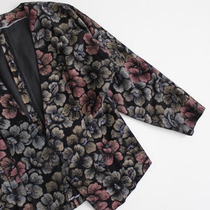 May include: A black floral patterned jacket with a long sleeve and a v-neck. The jacket has a subtle shimmer and a pink and gold floral pattern.