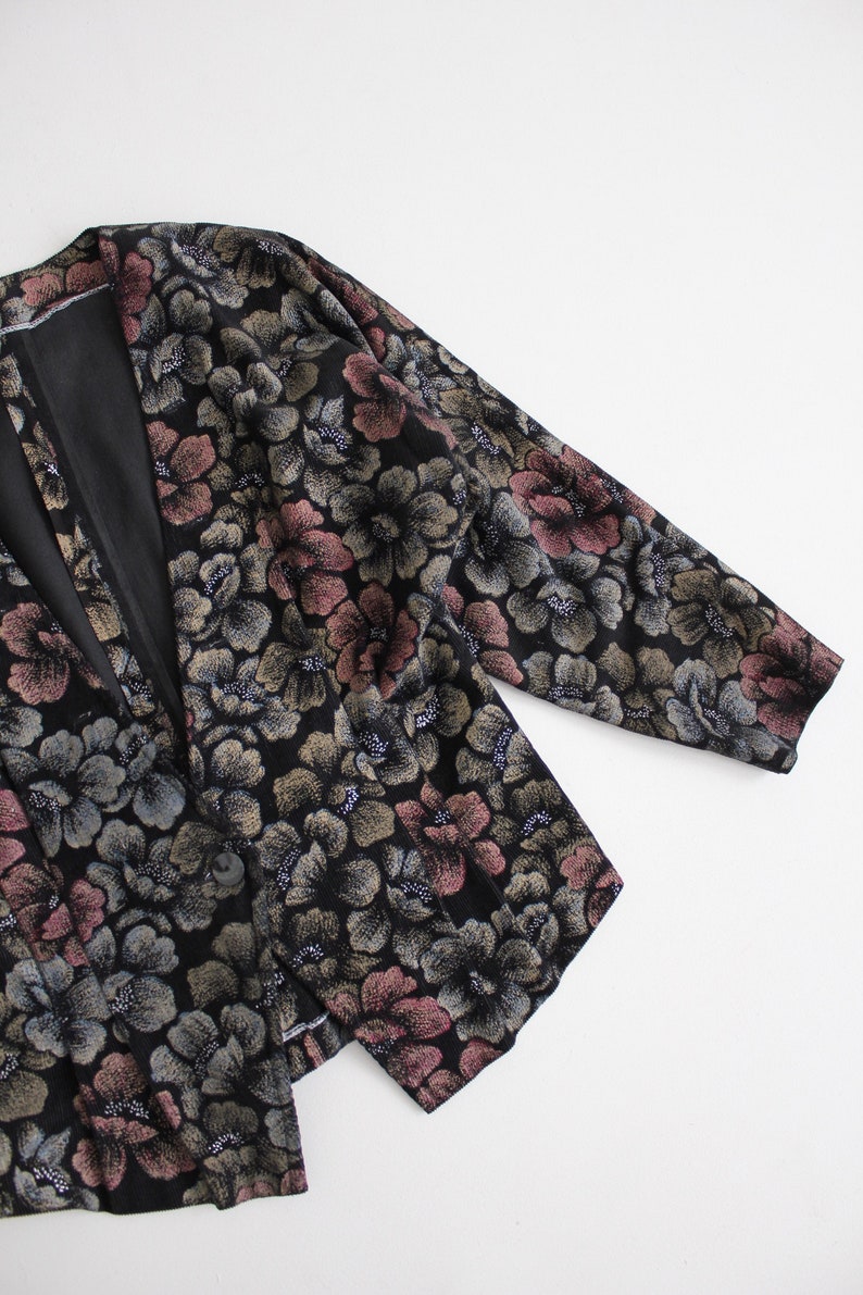 May include: A black floral patterned jacket with a long sleeve and a v-neck. The jacket has a subtle shimmer and a pink and gold floral pattern.