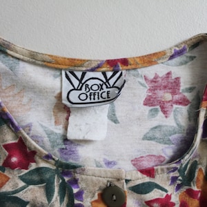May include: A beige button-up shirt with a floral print in shades of orange, red, purple, and green. The shirt has a label that reads "Box Office".