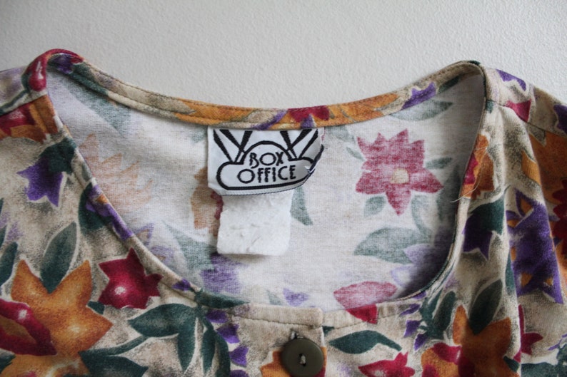 May include: A beige button-up shirt with a floral print in shades of orange, red, purple, and green. The shirt has a label that reads "Box Office".